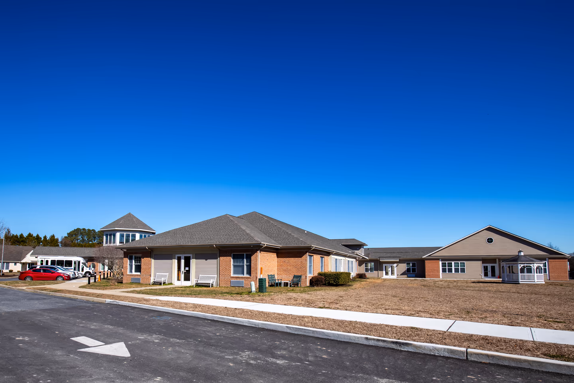Exterior view of Commonwealth Senior Living At Salisbury facility on a clear day, showing a single-story building with brick and siding, a paved driveway with a white directional arrow, parked vehicles, and a small white gazebo on the right side.