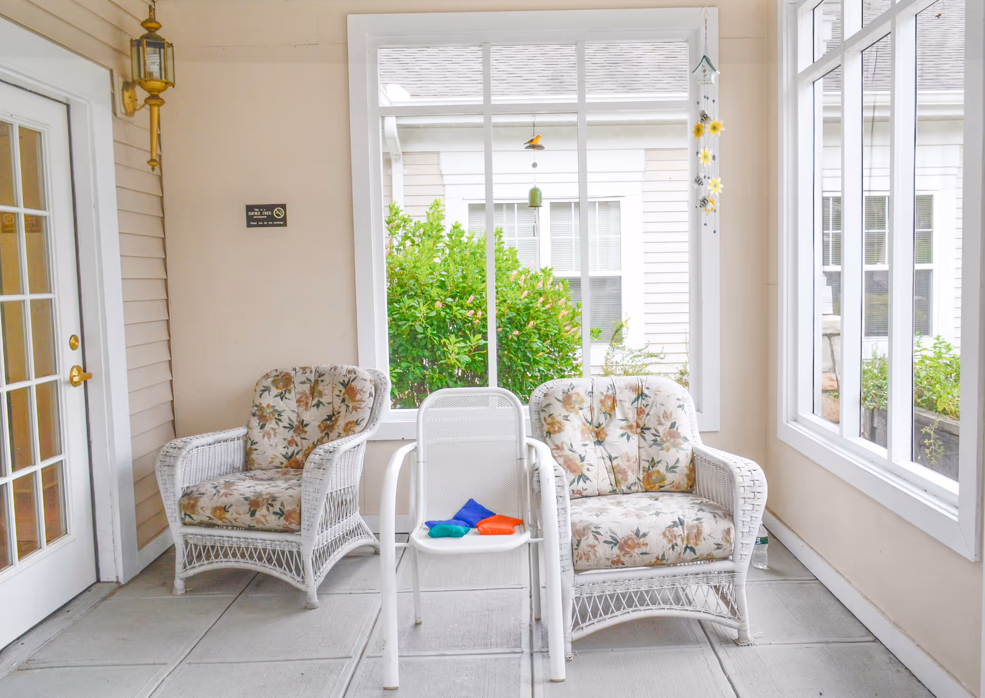 Enclosed sunroom-style seating area with two floral-pattern wicker armchairs, a white chair holding colorful beanbags, and large windows.