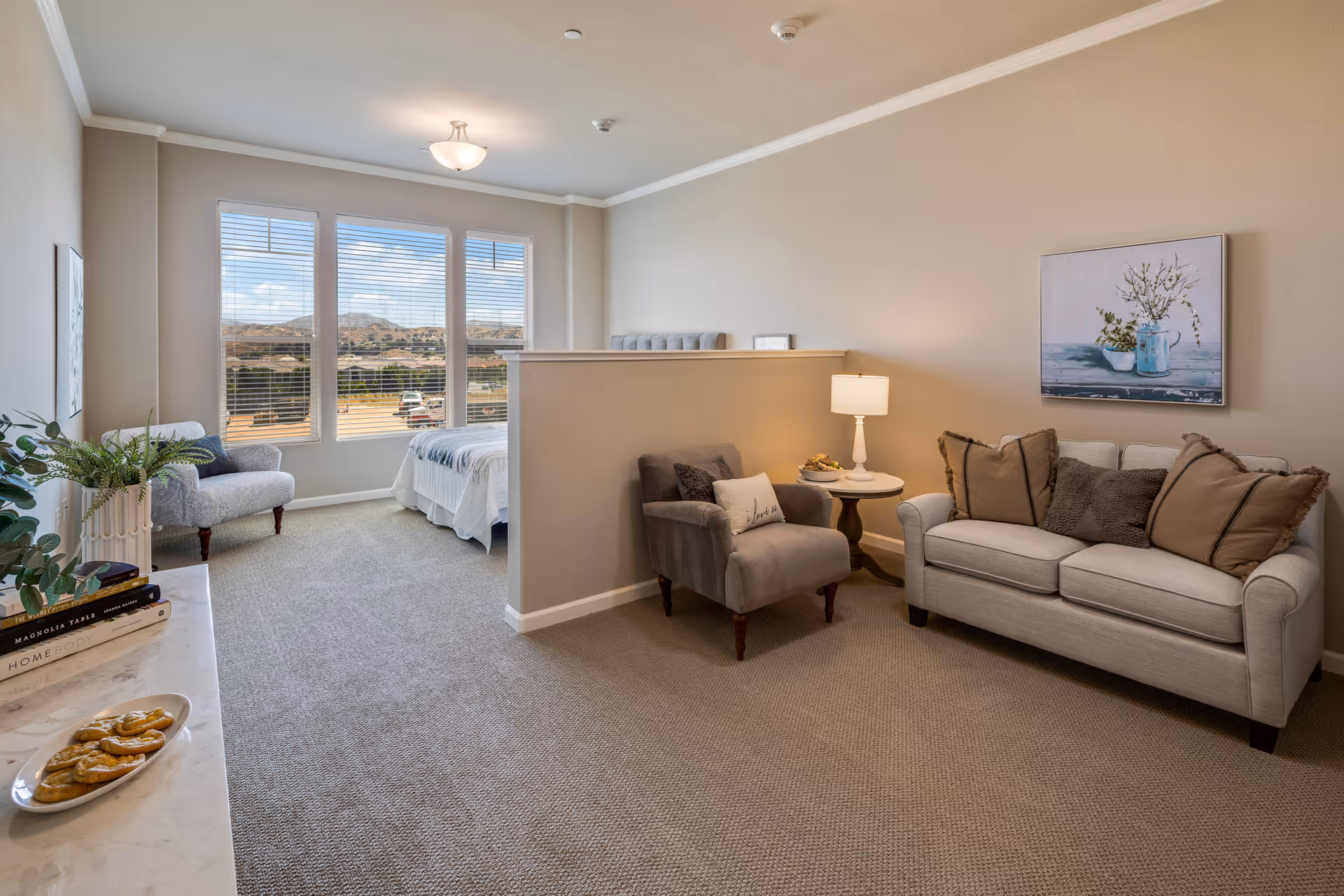 A cozy senior living suite with a sitting area and a sleeping area separated by a half wall. The sitting area features a light gray sofa with brown and dark gray pillows, a gray armchair with a pillow, a round side table with a lamp, and a painting of flowers on the wall. The sleeping area has a bed with white bedding and a gray headboard, next to large windows with blinds showing a view of hills and a parking lot outside. A marble countertop with a plate of cookies and some books is visible in the foreground.