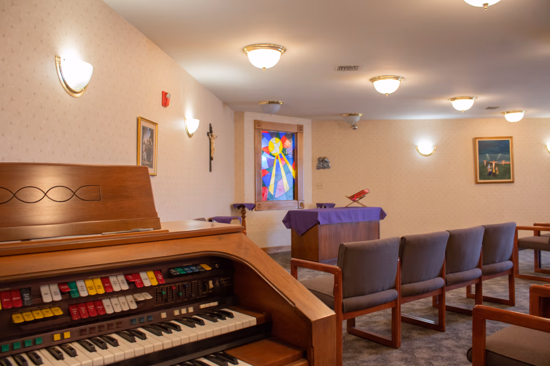 Interior view of a chapel or prayer room in a senior living facility featuring a wooden organ in the foreground, rows of cushioned chairs, a lectern draped with a purple cloth, a stained glass window, and religious artwork on the walls.