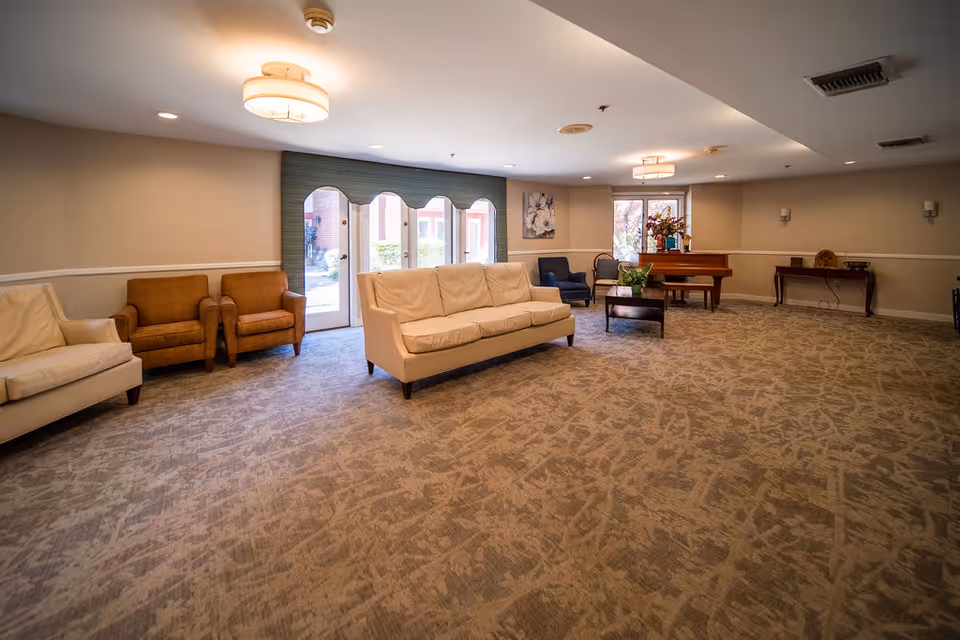 A spacious living room area with beige and brown armchairs, a cream-colored sofa, a coffee table with a plant, a piano with a flower arrangement on top, and a window letting in natural light. The room has patterned carpet flooring, neutral-colored walls, and ceiling lights.
