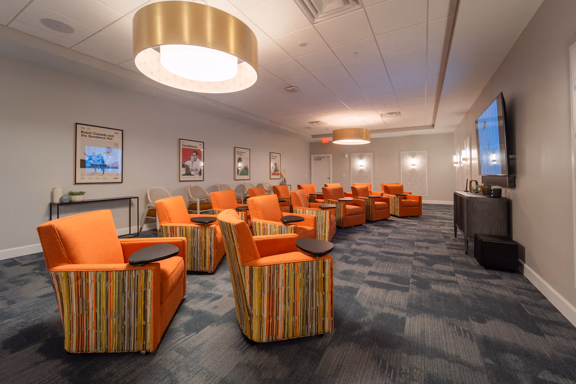 A modern lounge or common area with multiple orange armchairs featuring striped sides and attached black side tables, arranged facing a large flat-screen TV mounted on the wall. The room has a blue patterned carpet, light gray walls with framed movie posters, and two large circular ceiling lights providing warm illumination.