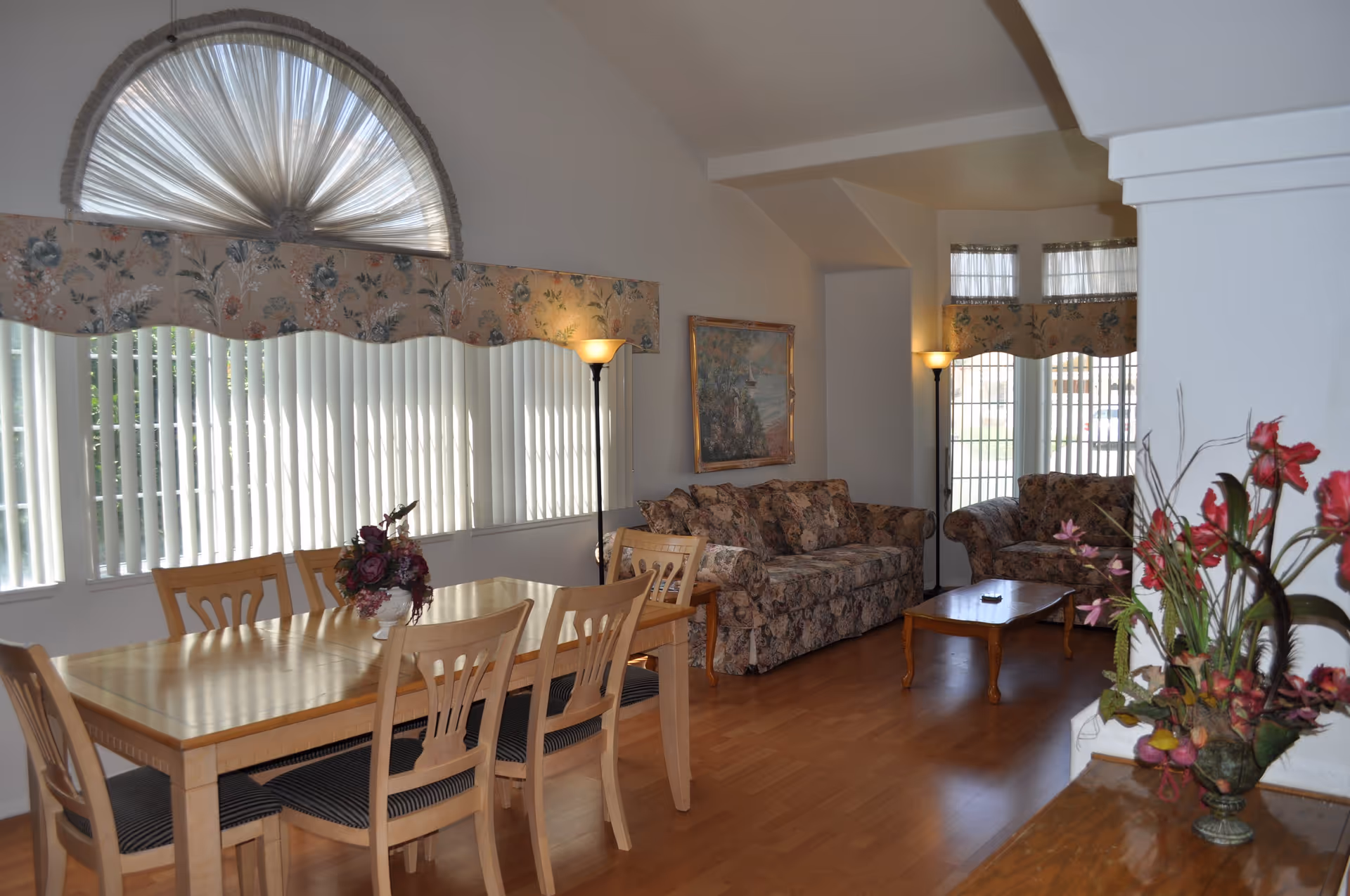 A combined dining and living room area with a wooden dining table and six chairs on the left, floral-patterned sofas and a wooden coffee table on the right, tall floor lamps, large windows with vertical blinds and floral valances, and a floral arrangement on a wooden surface in the foreground.