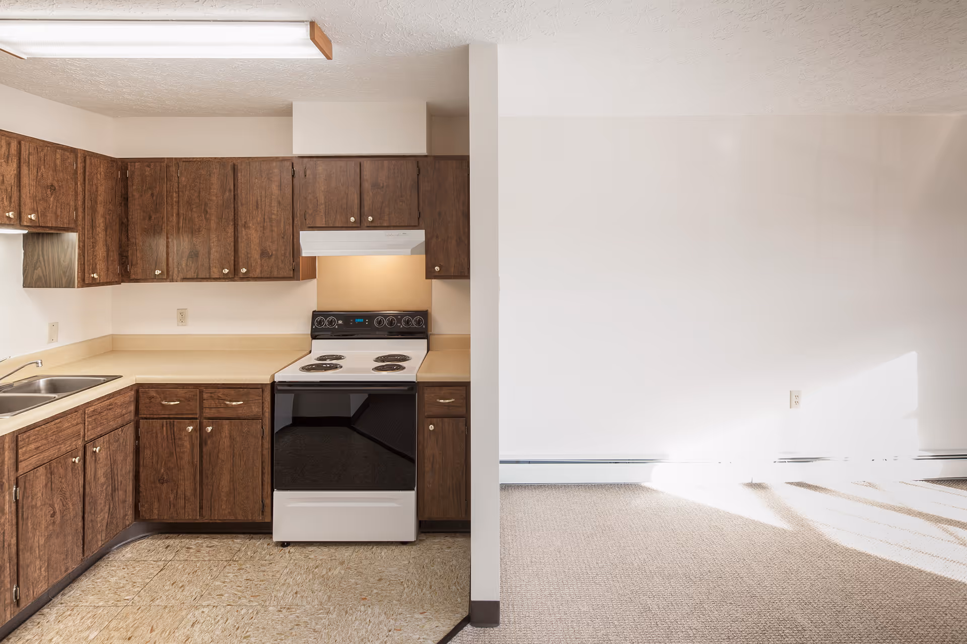 Small kitchen with dark wood cabinets, a sink, countertops and an electric stove next to a sunlit empty living area.