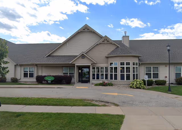 Front exterior view of a single-story building with beige siding and a gray shingled roof under a partly cloudy blue sky. The entrance has a small covered porch with glass doors, and there is a green sign near the entrance that reads 'Apple Creek Place'. There are bushes and flowers planted along the front of the building, and a sidewalk and driveway are visible in the foreground.
