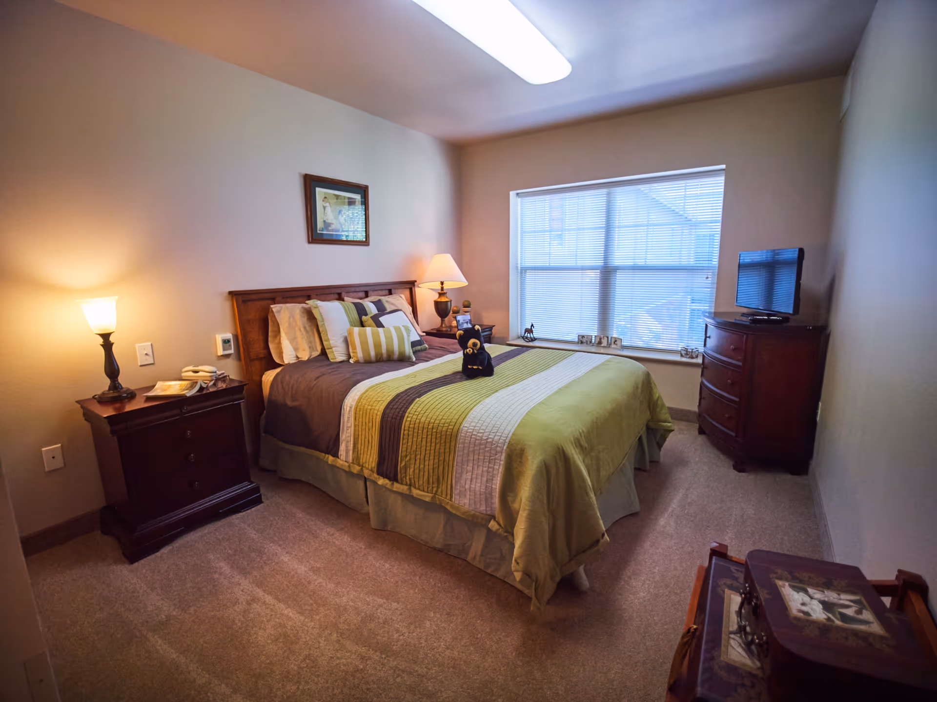Well-lit bedroom with a bed in green-striped bedding, nightstands with lamps, a dresser with TV, and a window with blinds.