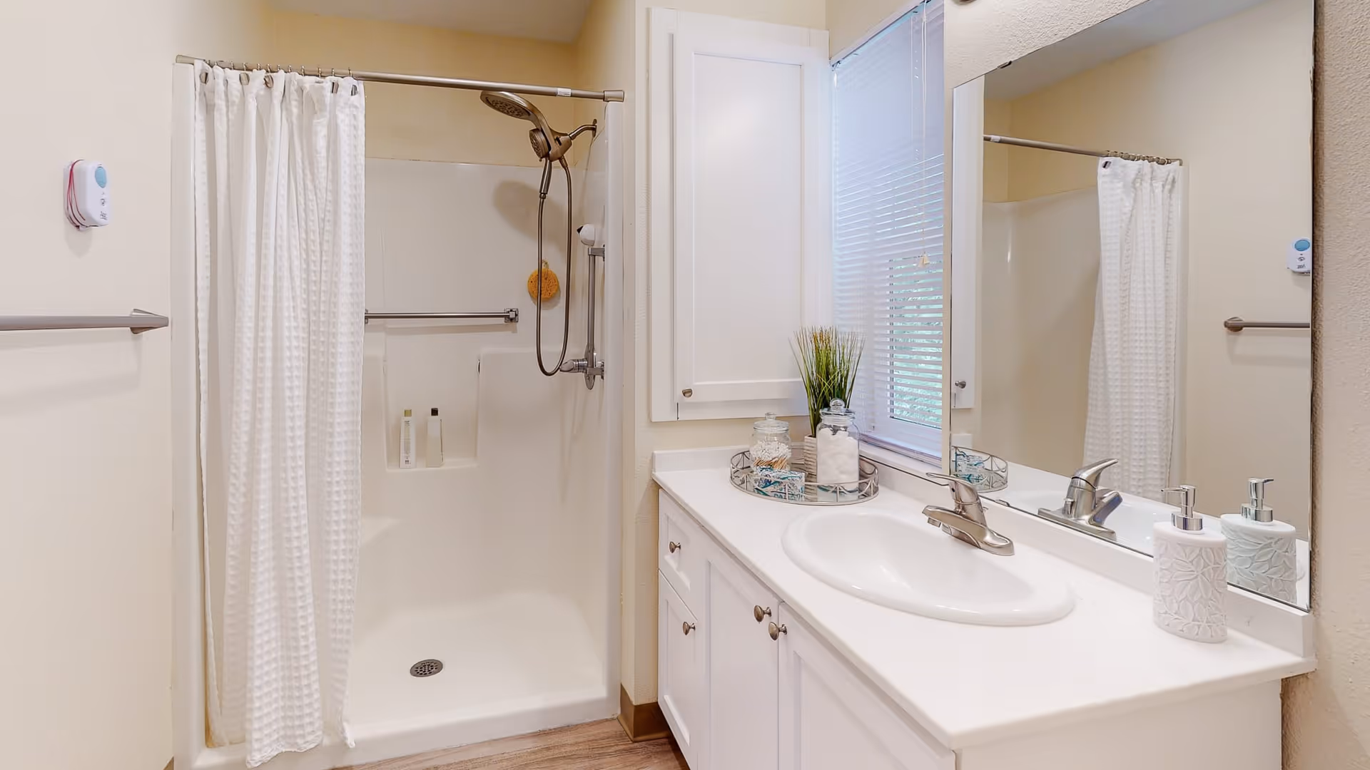 A clean and bright bathroom featuring a white shower with a curtain, a handheld showerhead, and built-in shelves holding toiletries. Next to the shower is a white vanity with a sink, a large mirror, a soap dispenser, and decorative items including a small plant and jars. A window with blinds is above the vanity.