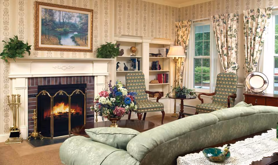 A cozy living room with a lit fireplace featuring a decorative screen. Above the fireplace is a framed painting of a serene landscape with a pond and trees. On either side of the fireplace are green plants. Two patterned armchairs with wooden arms are positioned near a window with floral curtains. A small round table with a lamp and a plant sits between the chairs. In the foreground, a green upholstered sofa with cushions faces the fireplace. A coffee table with a floral arrangement and decorative items is in front of the sofa. The walls have patterned wallpaper and built-in shelves with books and decorative items.