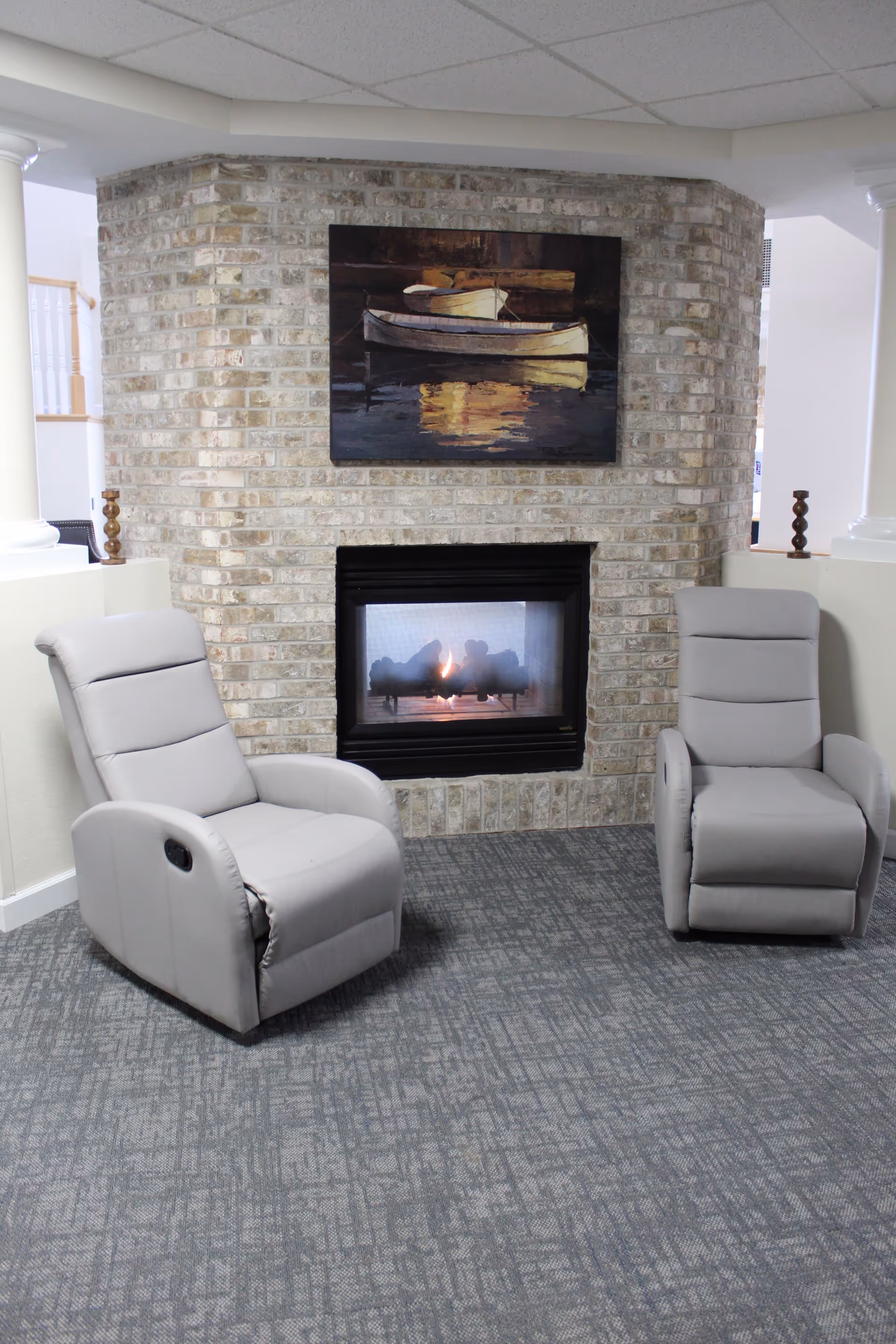 Two light gray recliner chairs positioned in front of a brick fireplace with a fire burning inside. Above the fireplace is a painting of two boats on water. The room has a gray patterned carpet and white walls with columns on either side.