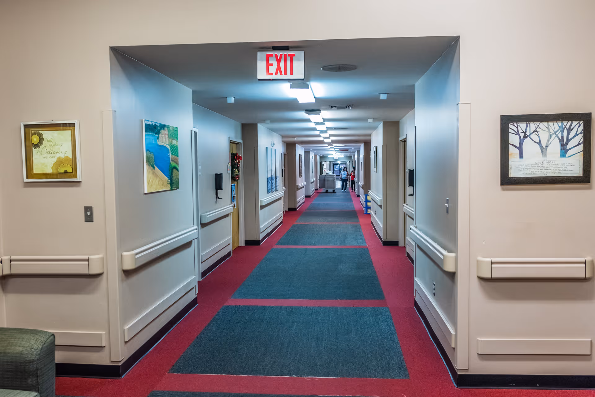 Long indoor hallway in a skilled nursing and rehab center with red and blue carpet, beige walls, handrails on both sides, framed artwork on the walls, and an exit sign overhead. Two people are visible in the distance at the end of the hallway.