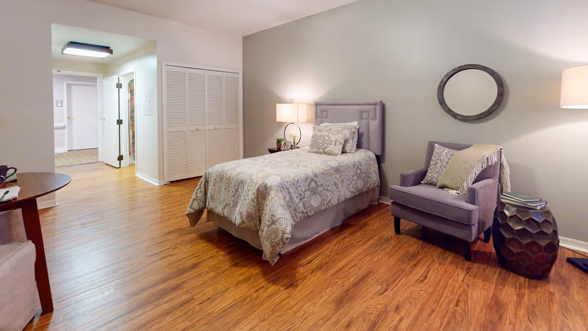 A cozy bedroom in a senior living facility with a single bed covered in a patterned bedspread, a bedside table with a lamp and small decor items, a comfortable armchair with pillows and a throw blanket, a small side table with books, a round mirror on the wall, and wooden flooring.