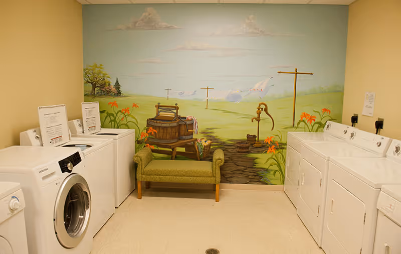 Laundry room with washers and dryers, a small green bench, and a painted pastoral mural on the back wall.
