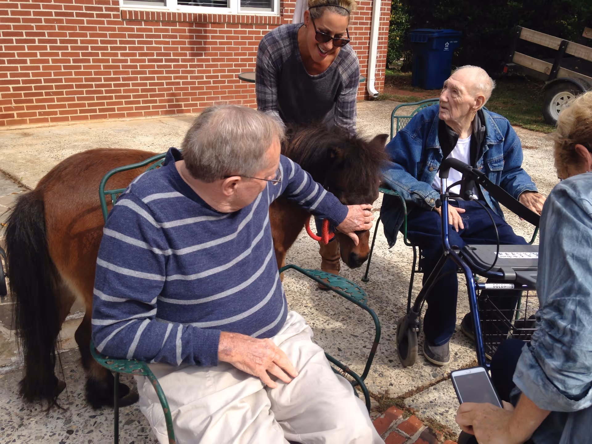 Two elderly men sitting outside on metal chairs, one petting a small brown pony, while a woman stands behind the pony smiling. Another person is seated nearby holding a smartphone. The setting is a paved patio area next to a brick building.