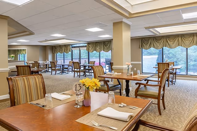 A spacious dining room with multiple wooden tables and chairs arranged neatly. Each table is set with placemats, napkins, utensils, and glasses. There are large windows with green valances allowing natural light to brighten the room. The carpeted floor and neutral-colored walls create a warm and inviting atmosphere.