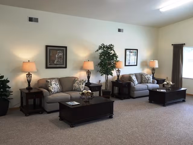 A spacious living room with two beige sofas each adorned with patterned throw pillows. Between the sofas are dark wooden side tables with matching table lamps. In front of each sofa is a dark wooden coffee table with decorative items. The room has light-colored walls, a large window with curtains on the right, and two framed artworks hanging on the wall. There are also two potted plants placed near the sofas.