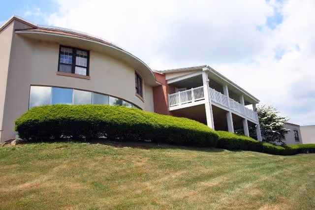 Exterior view of a senior living facility building with a curved wall and large windows, a white balcony with railings, and neatly trimmed green bushes in front on a grassy slope under a partly cloudy sky.