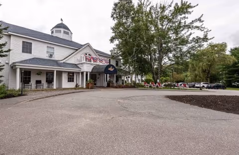 Front entrance of a white two-story senior living building with an awning, circular driveway, and surrounding trees.