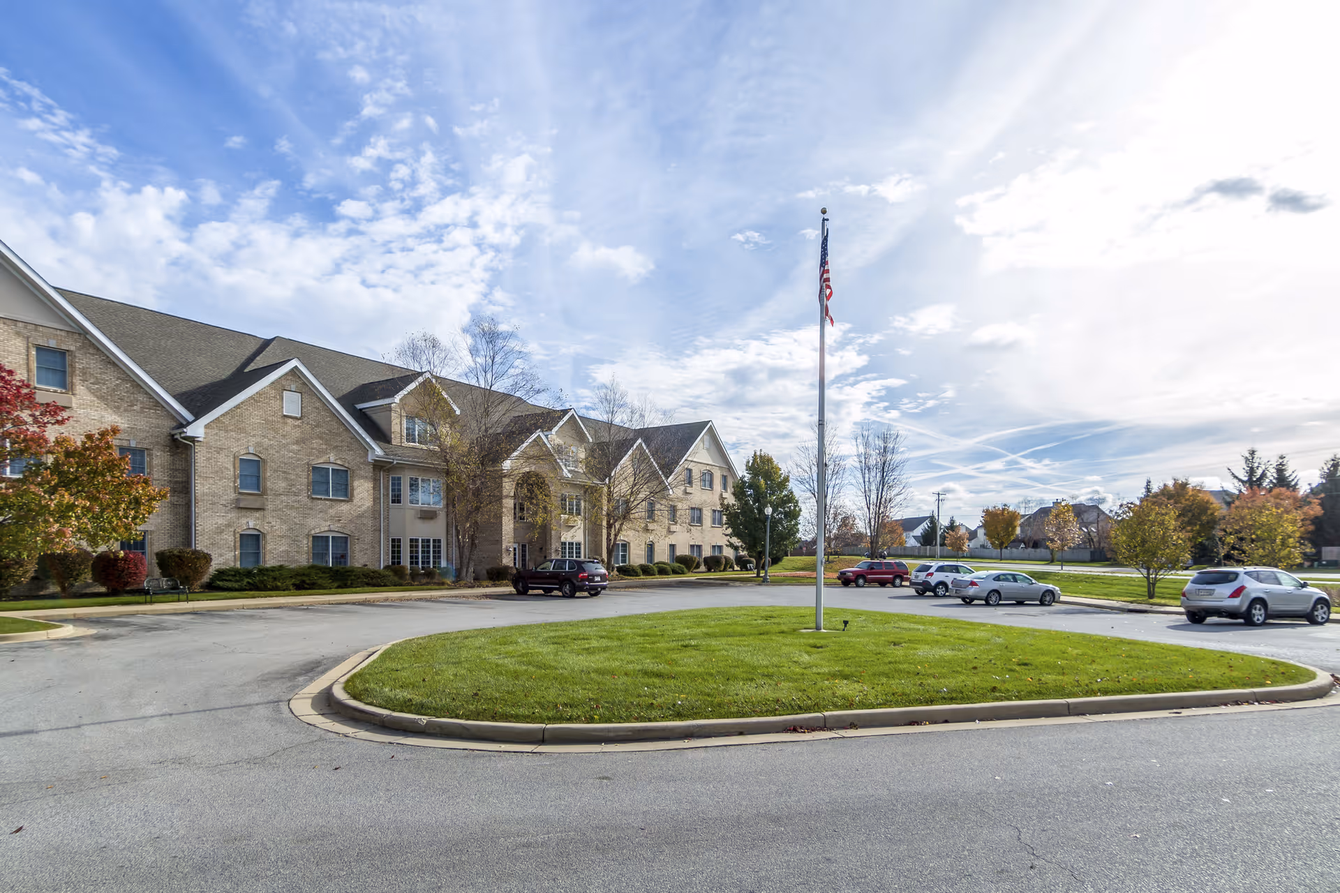 Brick assisted living building front with a circular driveway, flagpole, and parked cars under a partly cloudy sky.