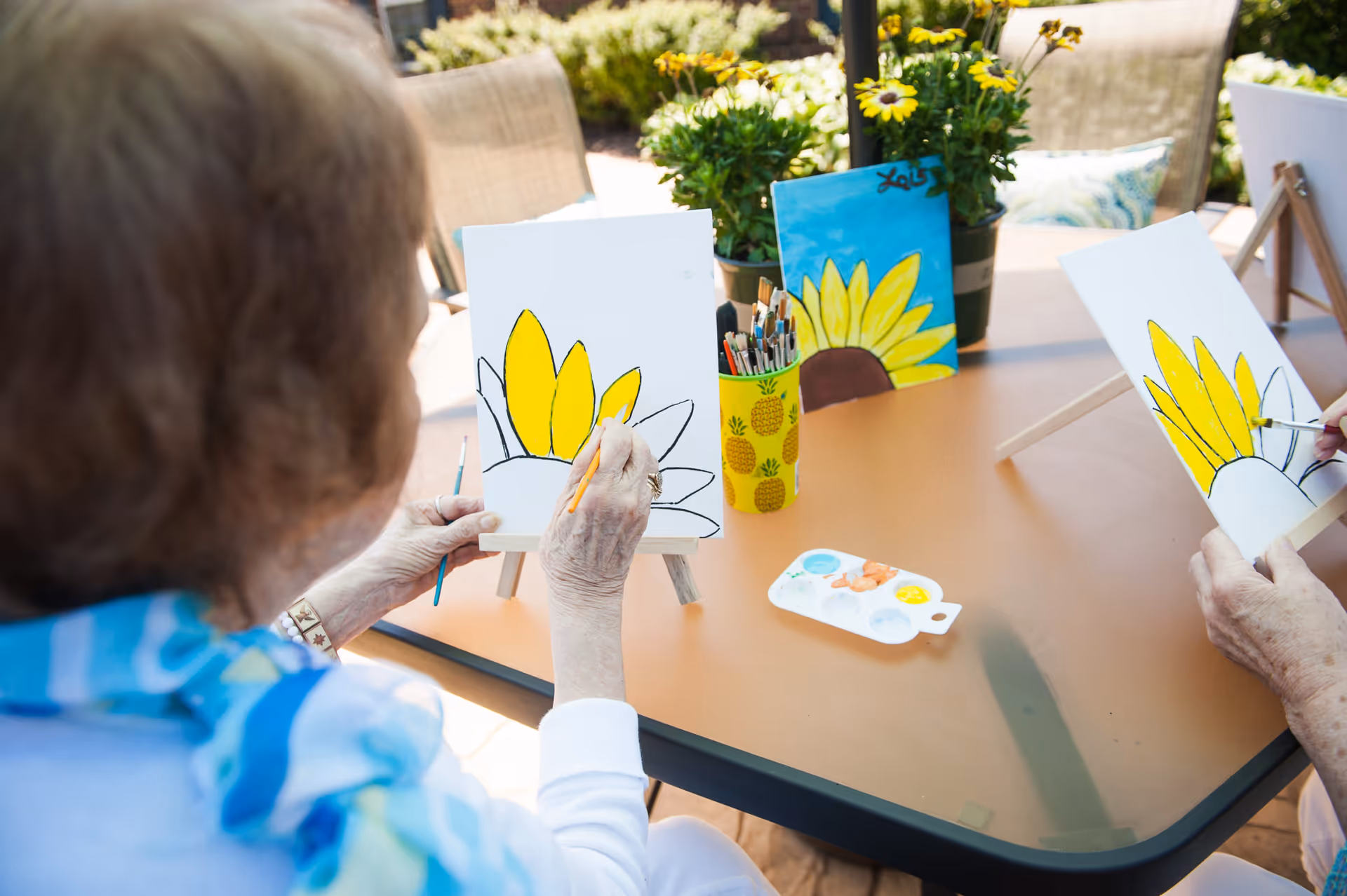 Two elderly individuals painting yellow sunflowers on canvases set on small easels at a table outdoors, with paintbrushes, a paint palette, and potted flowers on the table.