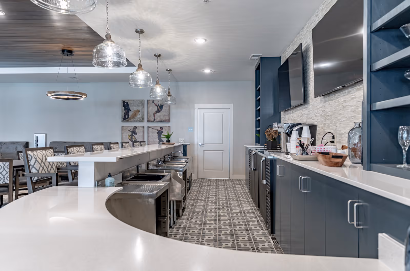 Modern communal kitchen and bar area with curved white countertops, pendant lights, bar seating, and built-in cabinets and TVs.