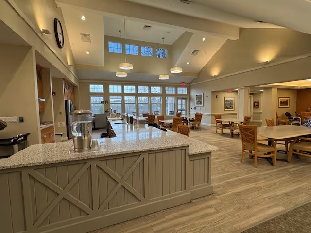 Spacious senior living dining and common area with a central service counter, multiple tables and chairs, large windows and a vaulted ceiling.