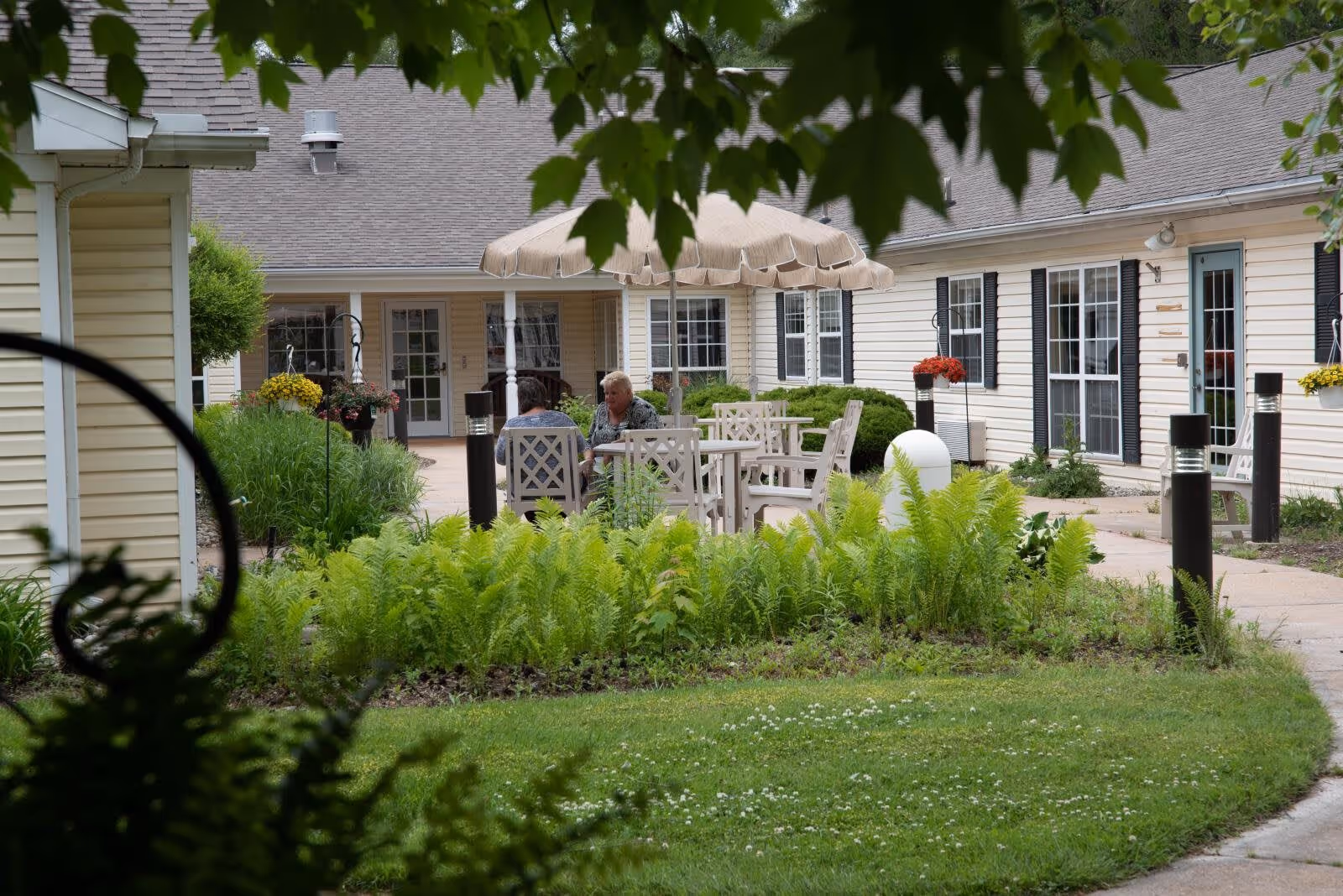 Two people sitting and talking at a white outdoor table with chairs under a beige umbrella in a garden courtyard area surrounded by green plants and shrubs, with beige buildings and windows in the background.