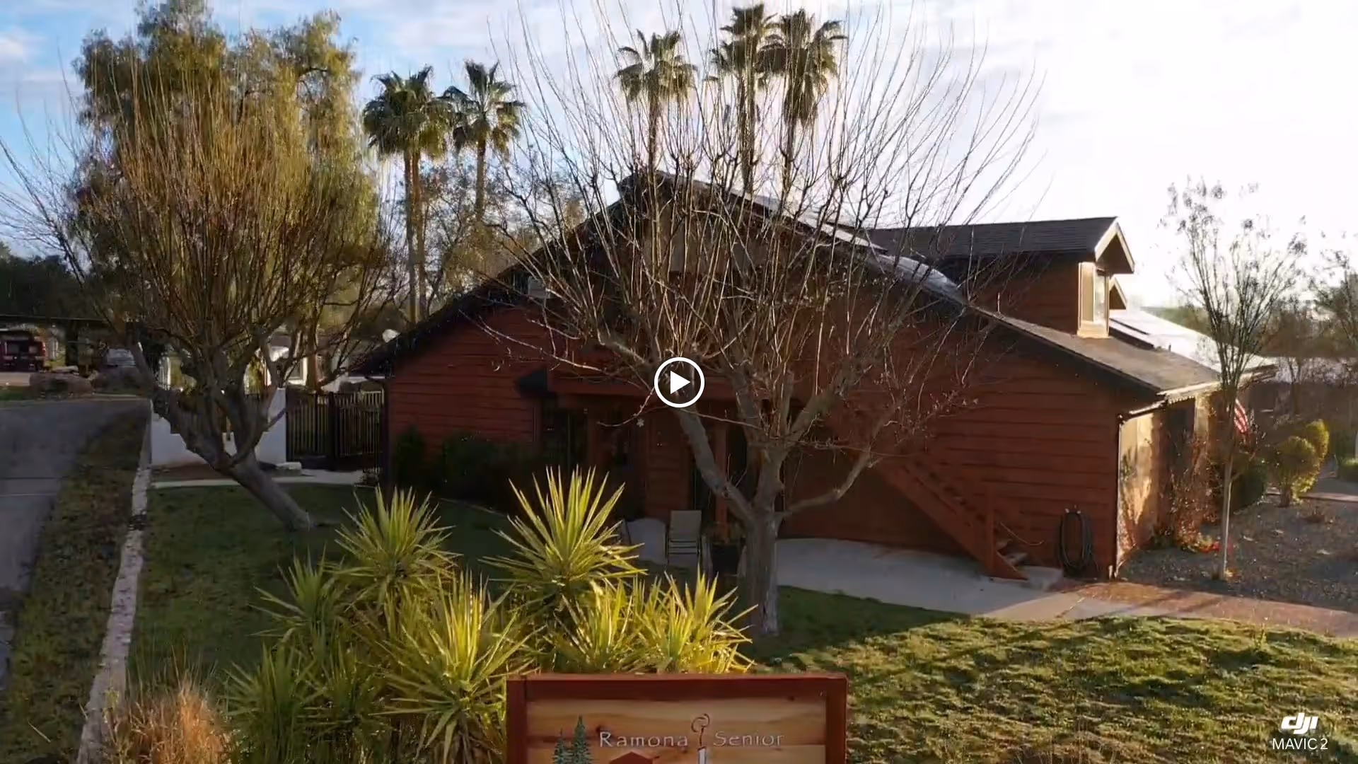 Exterior view of a single-story wooden building with a sloped roof, surrounded by trees and greenery under a partly cloudy sky. There is a sign in front that reads 'Ramona Senior'.