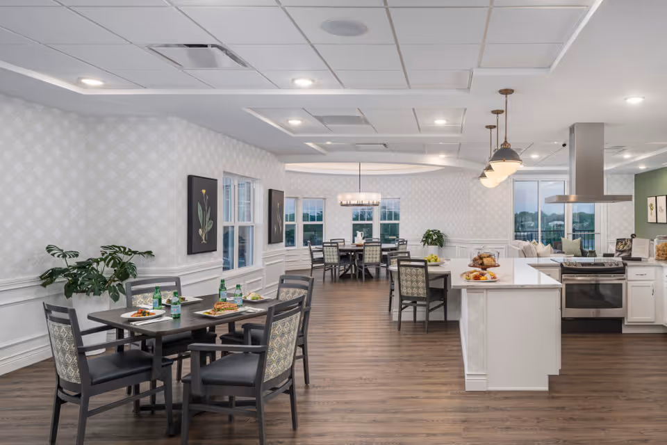 A bright and spacious dining and kitchen area in a senior living community. The room features multiple dining tables with chairs, set with plates of food and bottled drinks. The kitchen area includes a large island with a stove, oven, and hanging pendant lights. The space has wood flooring, patterned wallpaper, and large windows letting in natural light.