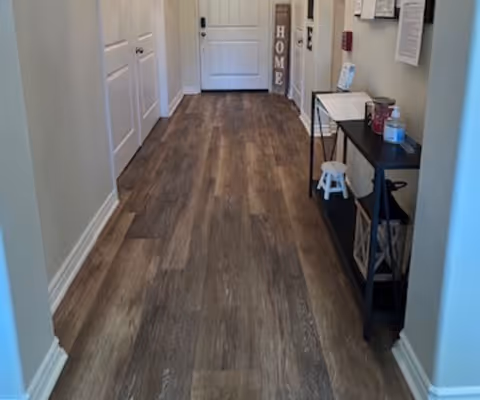 A hallway with wood-look flooring, white walls, and several white doors on the left side. On the right side, there is a black console table with various items including hand sanitizer and a small white stool underneath. At the end of the hallway, a decorative sign with the word 'HOME' is visible next to a closed white door.