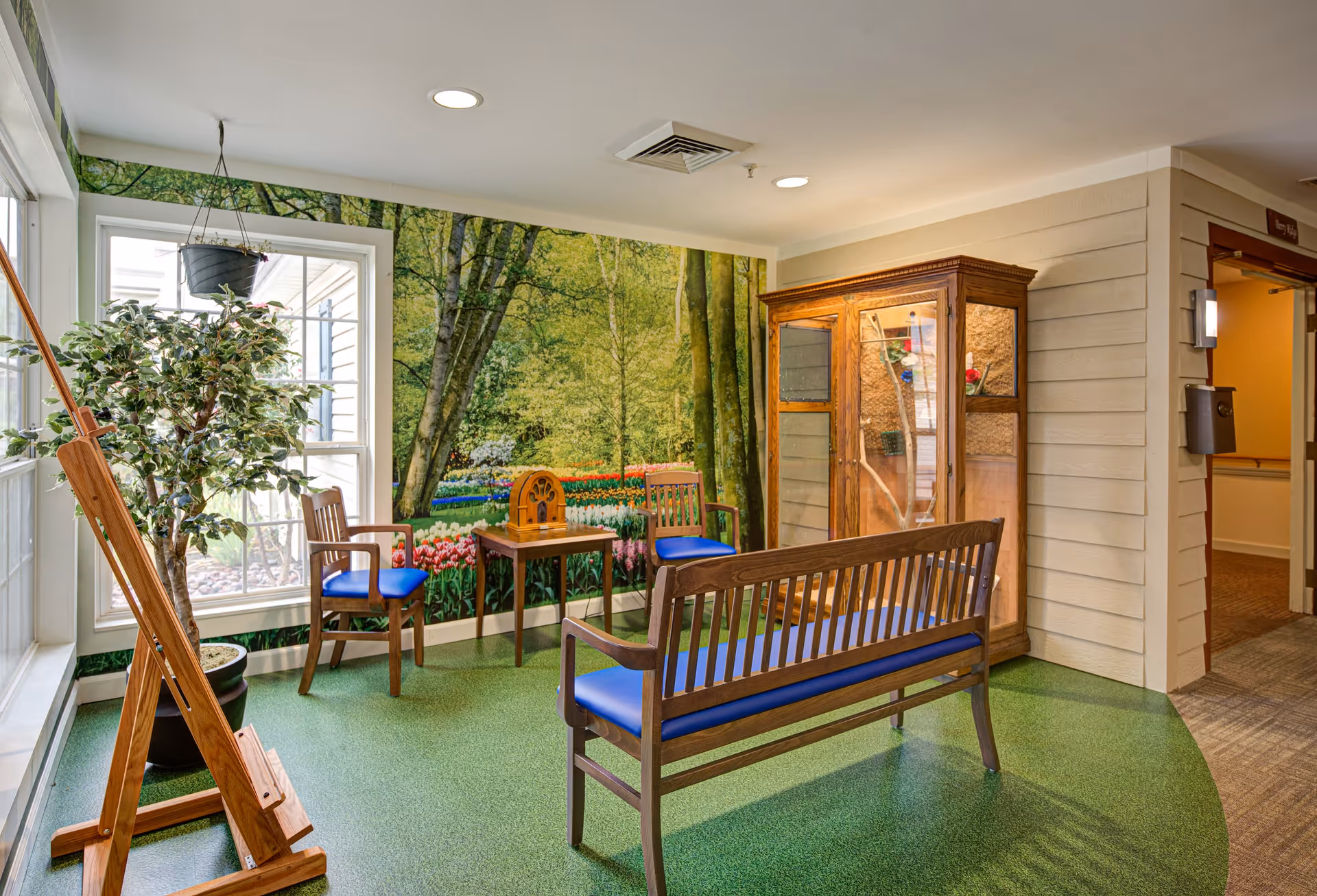 A cozy indoor seating area with wooden chairs and a bench featuring blue cushions, a small wooden table with a vintage radio, a large potted plant, and an easel. The back wall has a large mural depicting a lush garden with trees and colorful flowers, creating a serene atmosphere. There is also a wooden display cabinet and a hallway entrance to the right.