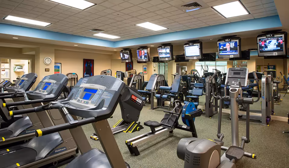 Interior view of a fitness center with multiple exercise machines including treadmills, stationary bikes, and weight machines. Several televisions are mounted on the wall above the equipment, displaying various channels. The room has a drop ceiling with fluorescent lighting and mirrors on the far wall.