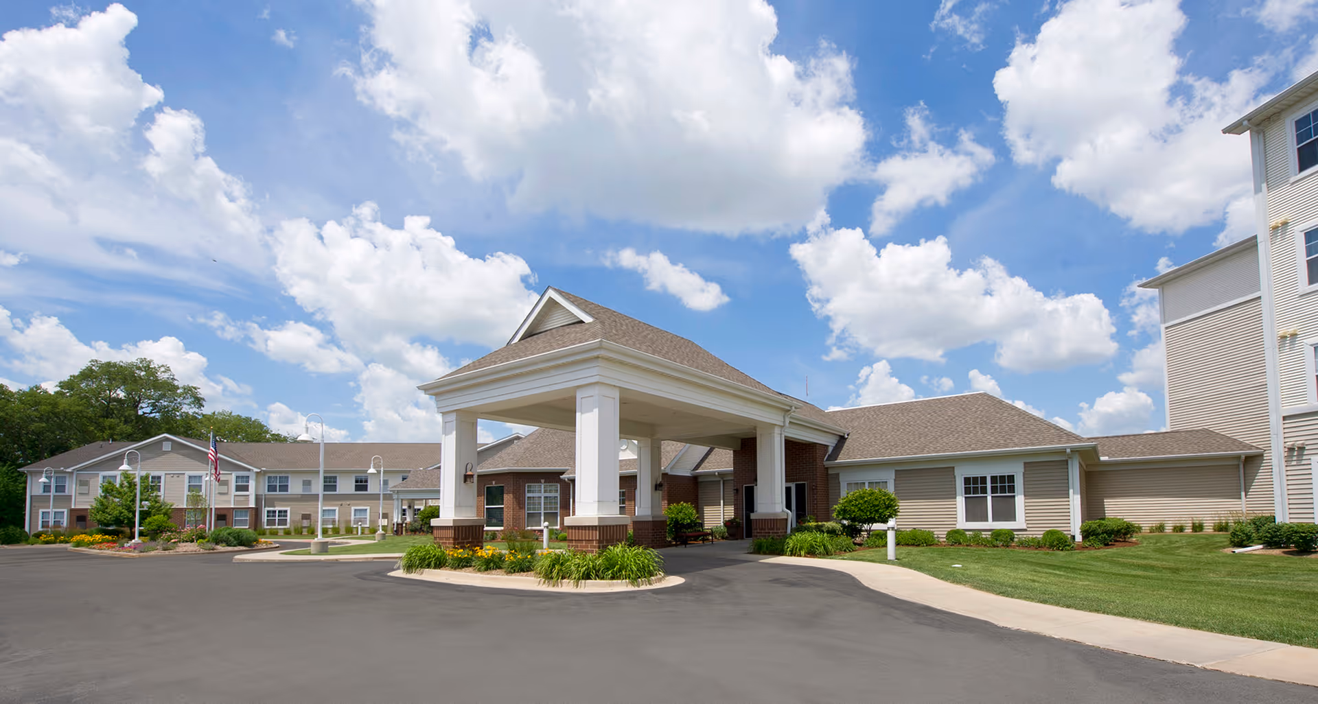 Front entrance of a senior living community with a covered porte-cochere, landscaped grounds, and a blue sky with clouds.