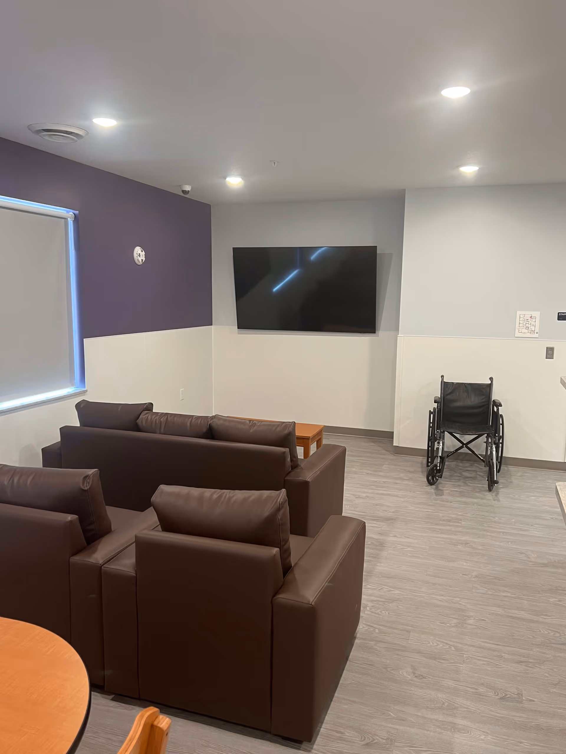 A small lounge area with two brown leather sofas facing a wall-mounted flat screen TV. There is a wooden coffee table between the sofas. A wheelchair is positioned near the wall on the right side. The walls are painted white and purple, and the floor has a light wood finish. Ceiling lights illuminate the room.