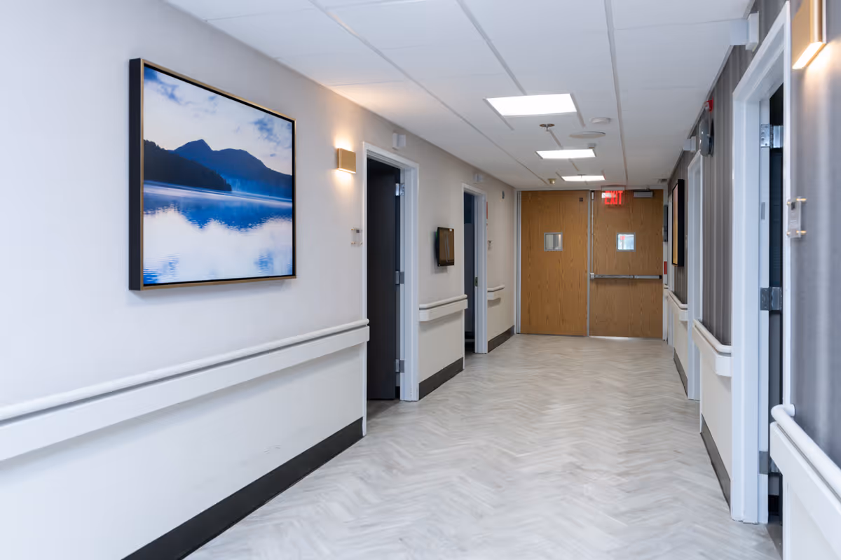 A clean, well-lit hallway in a healthcare facility with light-colored walls and flooring. The hallway features handrails on both sides, several open doorways, a large framed picture of a serene lake and mountain scene on the left wall, and double wooden doors at the far end with small windows and an illuminated exit sign above.