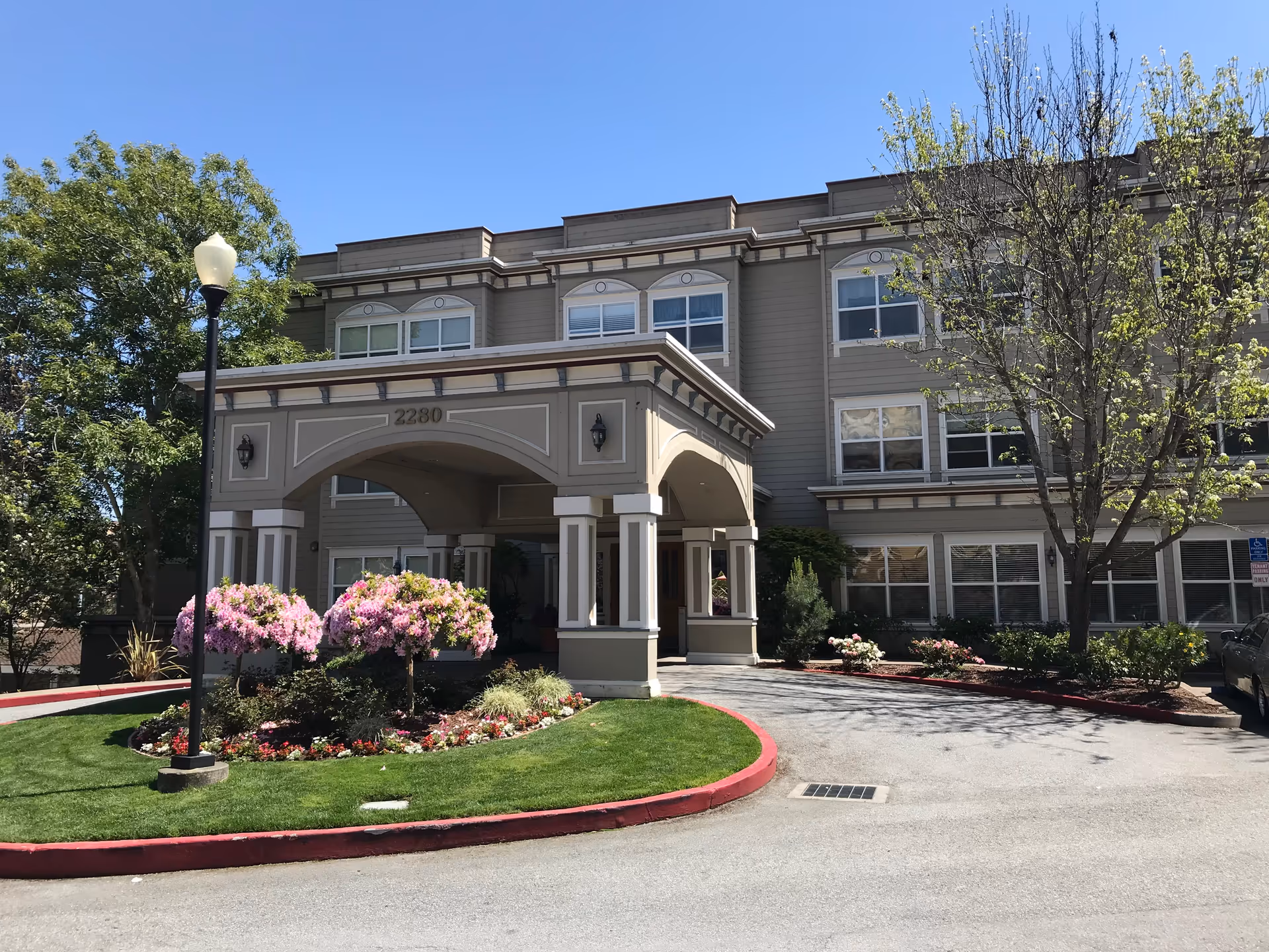 Exterior view of a three-story senior living facility building with a covered entrance. The building is beige with white trim and has multiple windows. There is a landscaped area with green grass, pink flowering bushes, and a street lamp near the entrance. The sky is clear and blue.