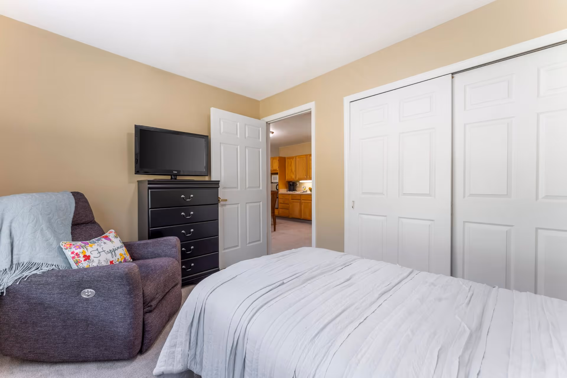 A bedroom with a bed covered in a white comforter, a dark gray armchair with a light blue throw blanket and a floral pillow, a black chest of drawers with a flat-screen TV on top, beige walls, and white closet doors. The bedroom door is open, showing a view into a kitchen area with wooden cabinets.