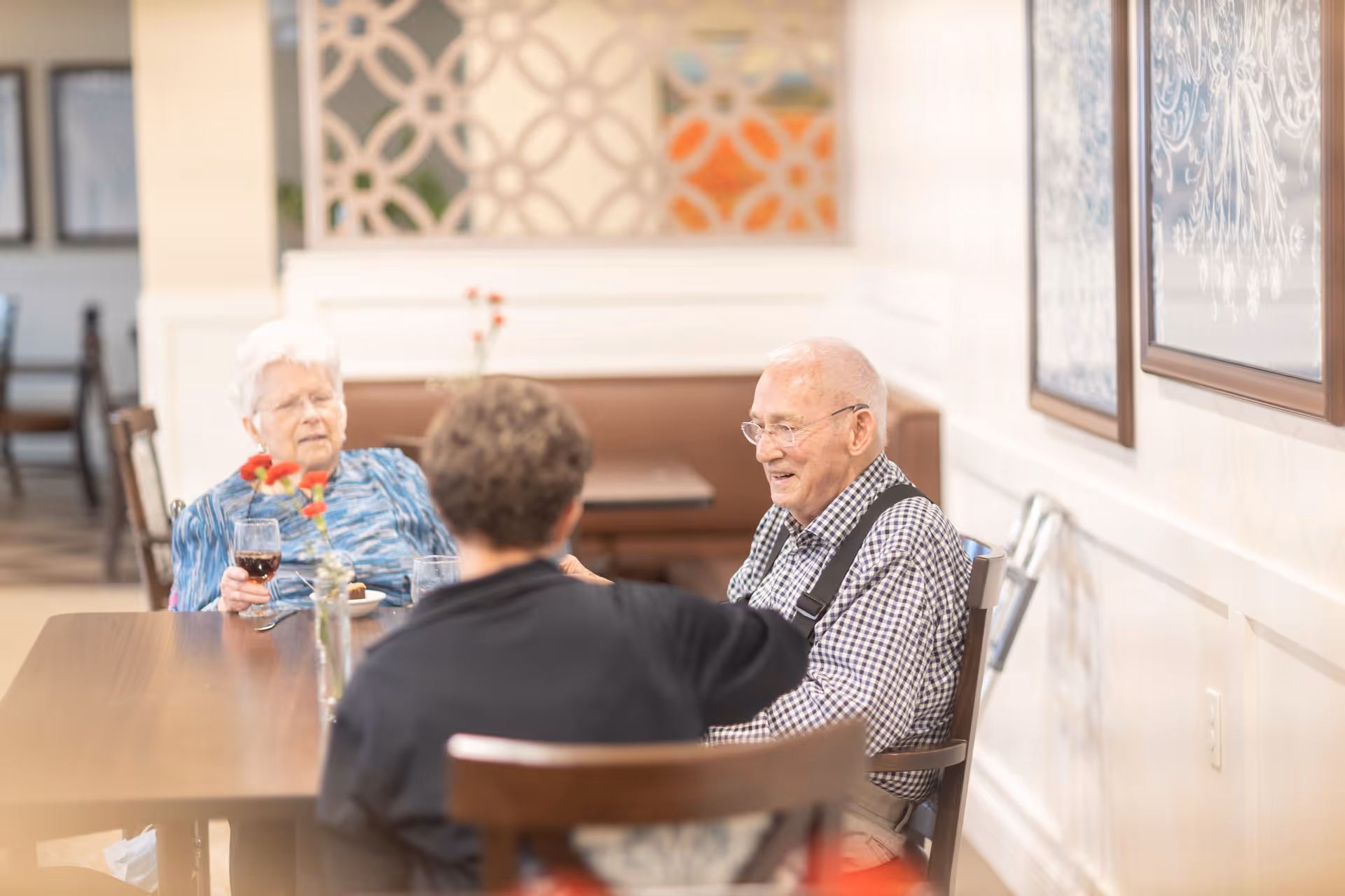 Three elderly people sitting around a wooden table in a well-lit dining area, engaging in conversation. One woman holds a glass of red wine, and there are flowers in a vase on the table. The background shows decorative wall panels and framed artwork.