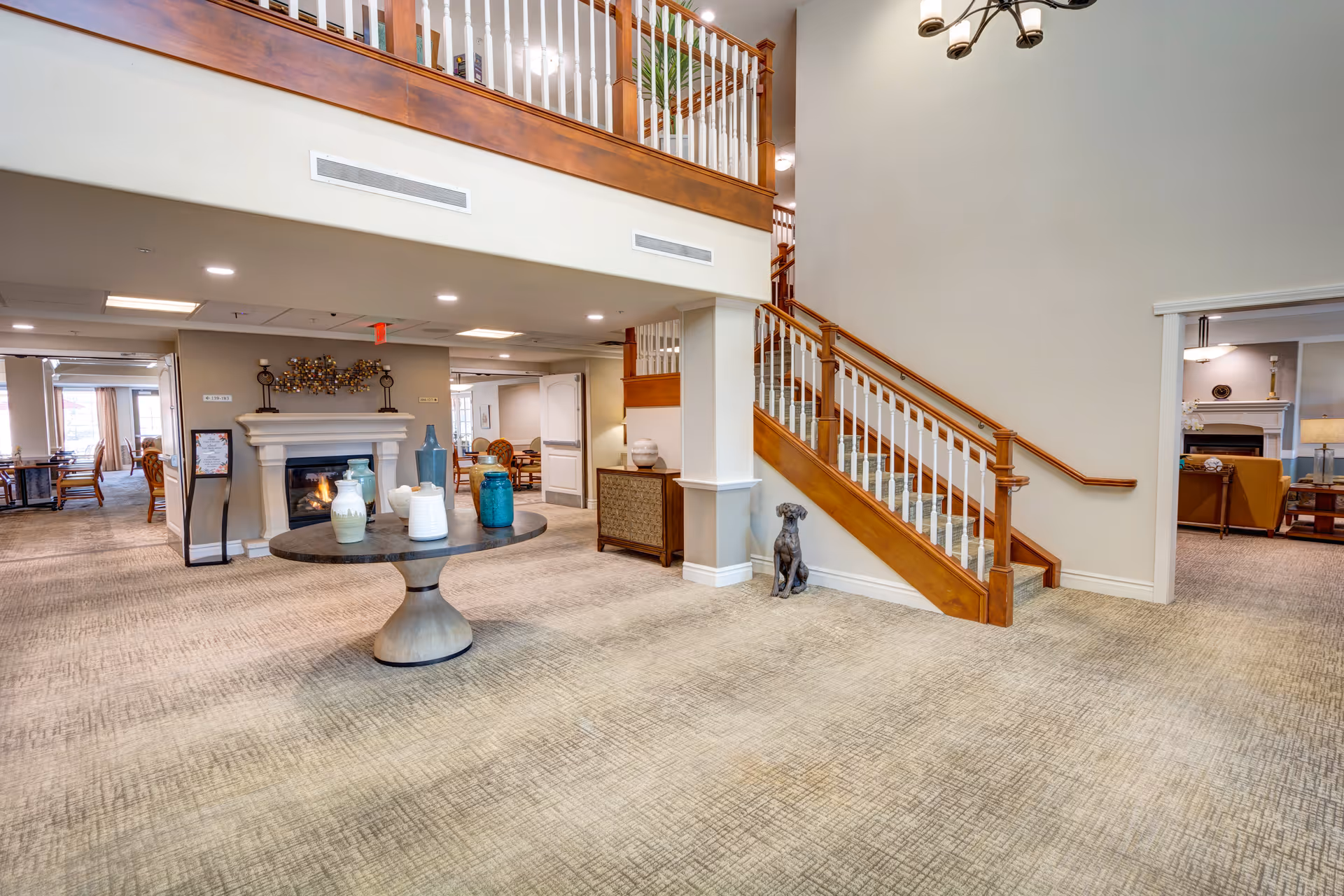 Spacious interior area of a senior living facility featuring a carpeted floor, a round table with decorative vases, a staircase with wooden handrails and white balusters, a fireplace with decorative items above it, and seating areas visible in adjoining rooms.