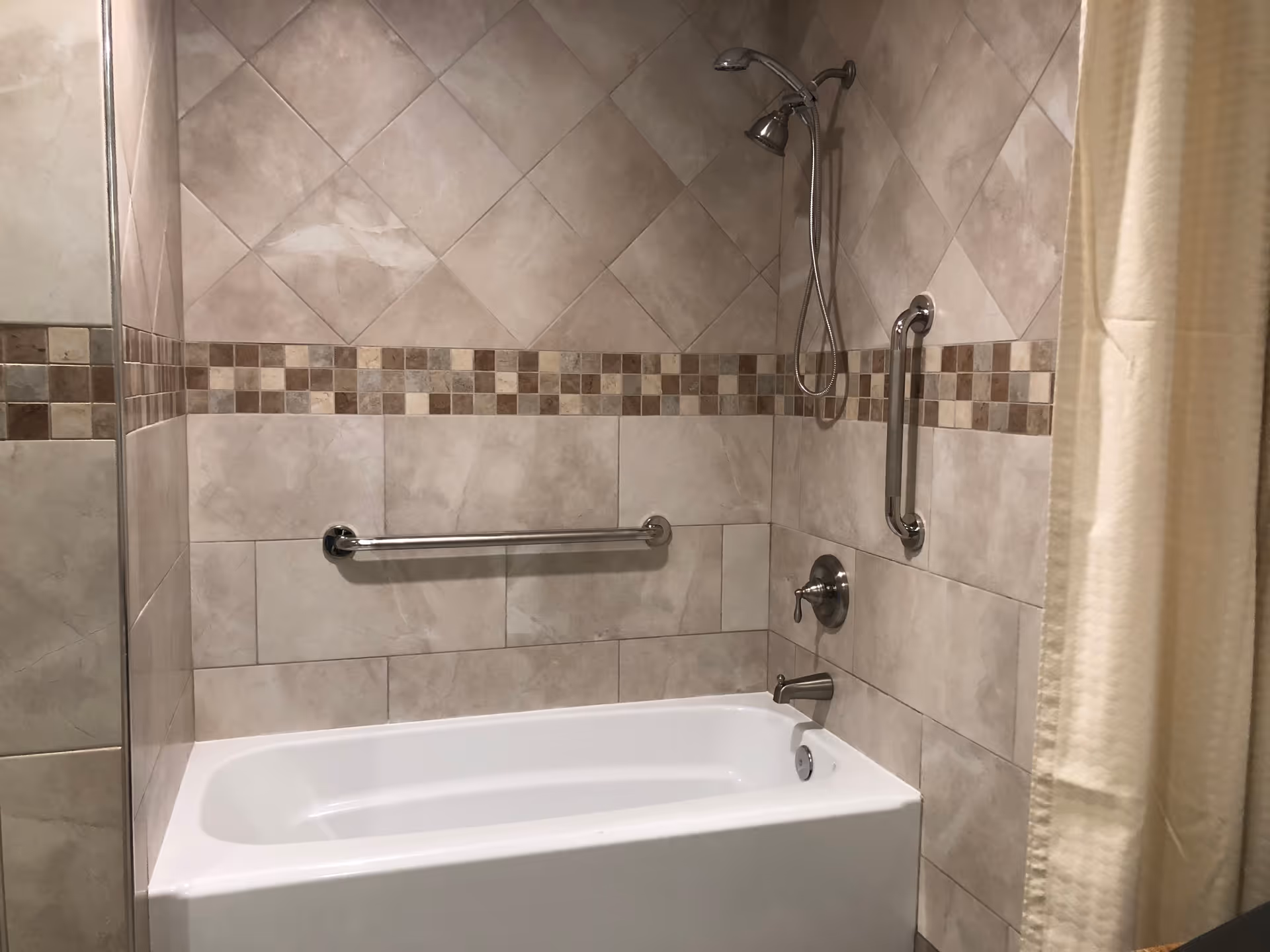 A bathroom with a white bathtub surrounded by beige and brown tiled walls. The tiles include a decorative strip of small square tiles in various shades of brown. There is a silver showerhead, a faucet, and two silver grab bars mounted on the tiled walls. A beige shower curtain is partially visible on the right side.