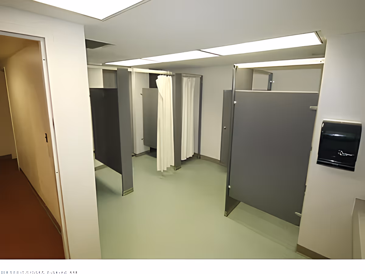 Interior view of a communal bathroom area with multiple shower stalls separated by gray partitions and white shower curtains. The floor is light green, and the walls are white. There is a paper towel dispenser mounted on the wall to the right.