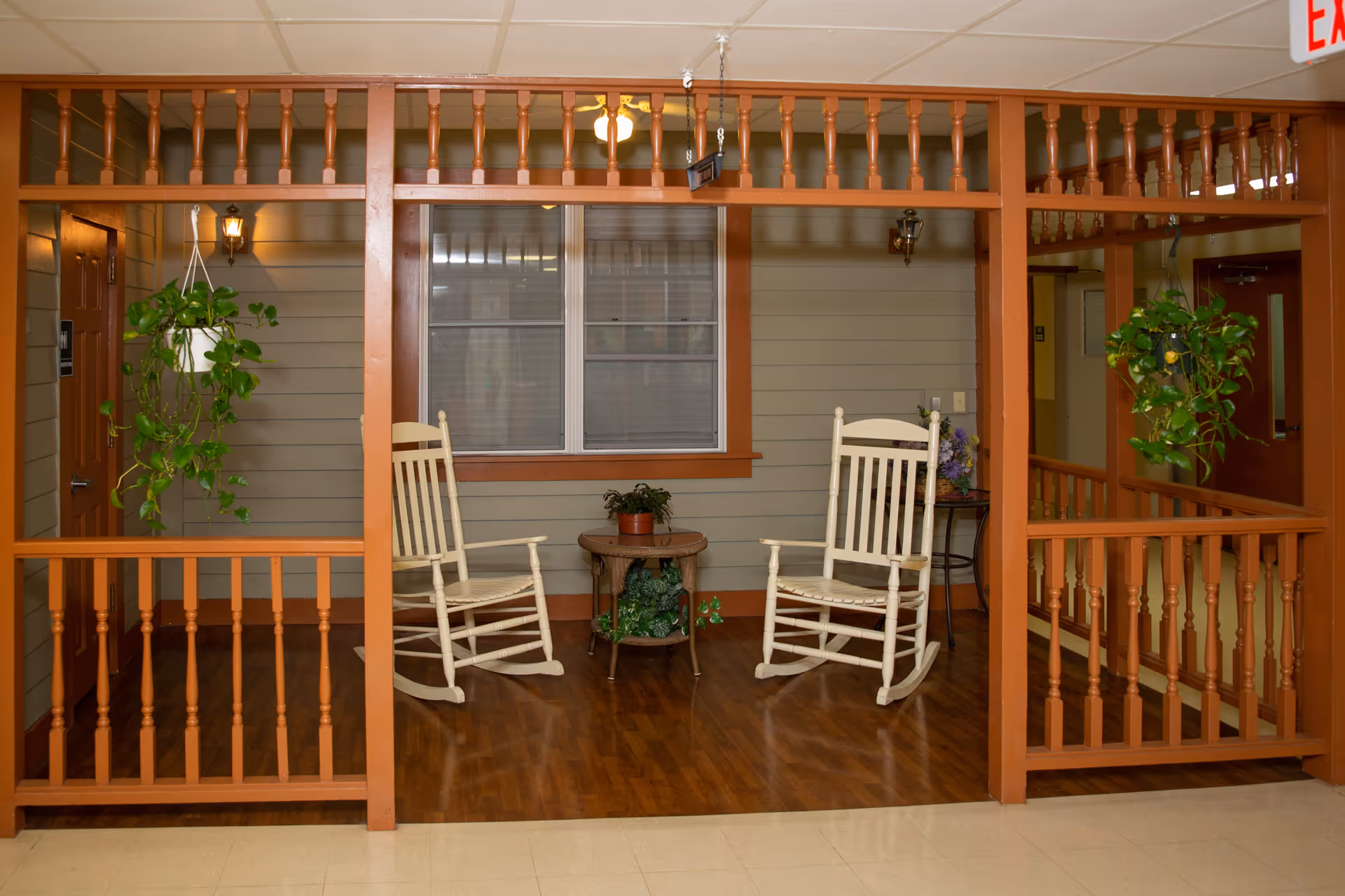 Indoor sitting area with two white wooden rocking chairs facing each other, a small round table with a potted plant between them, hanging green plants on either side, and a window with blinds in the background. The area is framed by wooden railings and posts, with beige walls and a wooden floor.