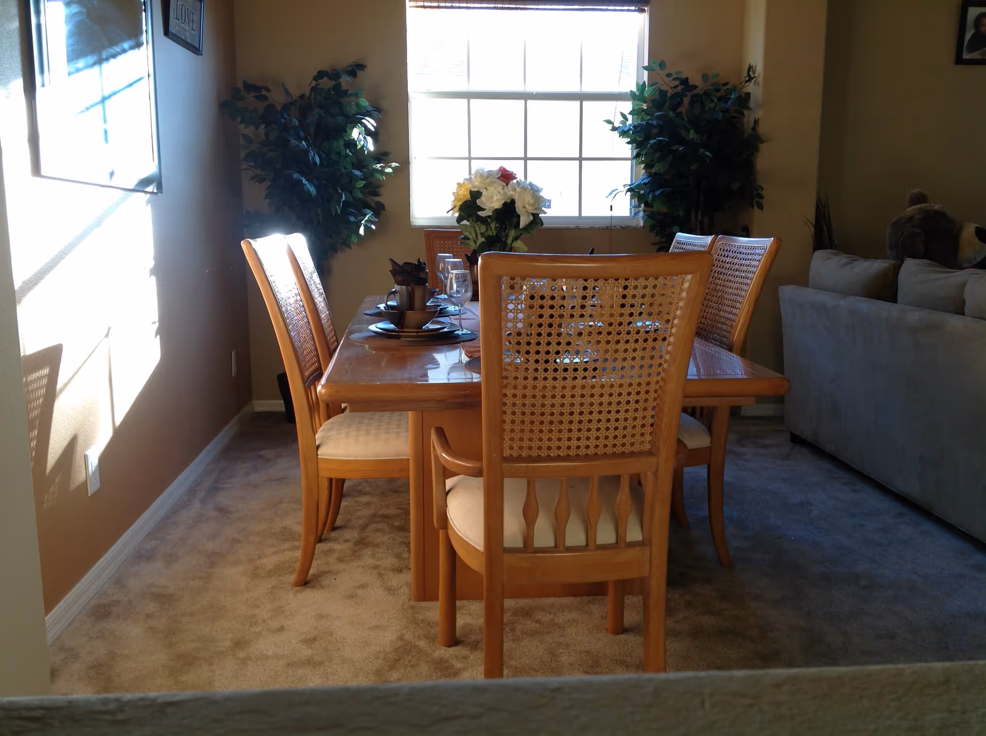 A dining area with a wooden table and six matching chairs with woven backs. The table is set with plates, bowls, cups, and wine glasses, and a vase with white and red flowers is placed in the center. Two large green potted plants are positioned near a window with white blinds, allowing natural light to fill the room. A beige carpet covers the floor, and part of a gray sofa is visible to the right.