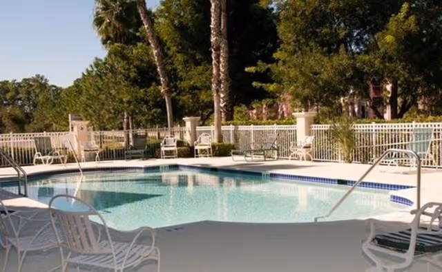 Outdoor swimming pool surrounded by lounge chairs, white fencing, and palm trees at a senior living facility.