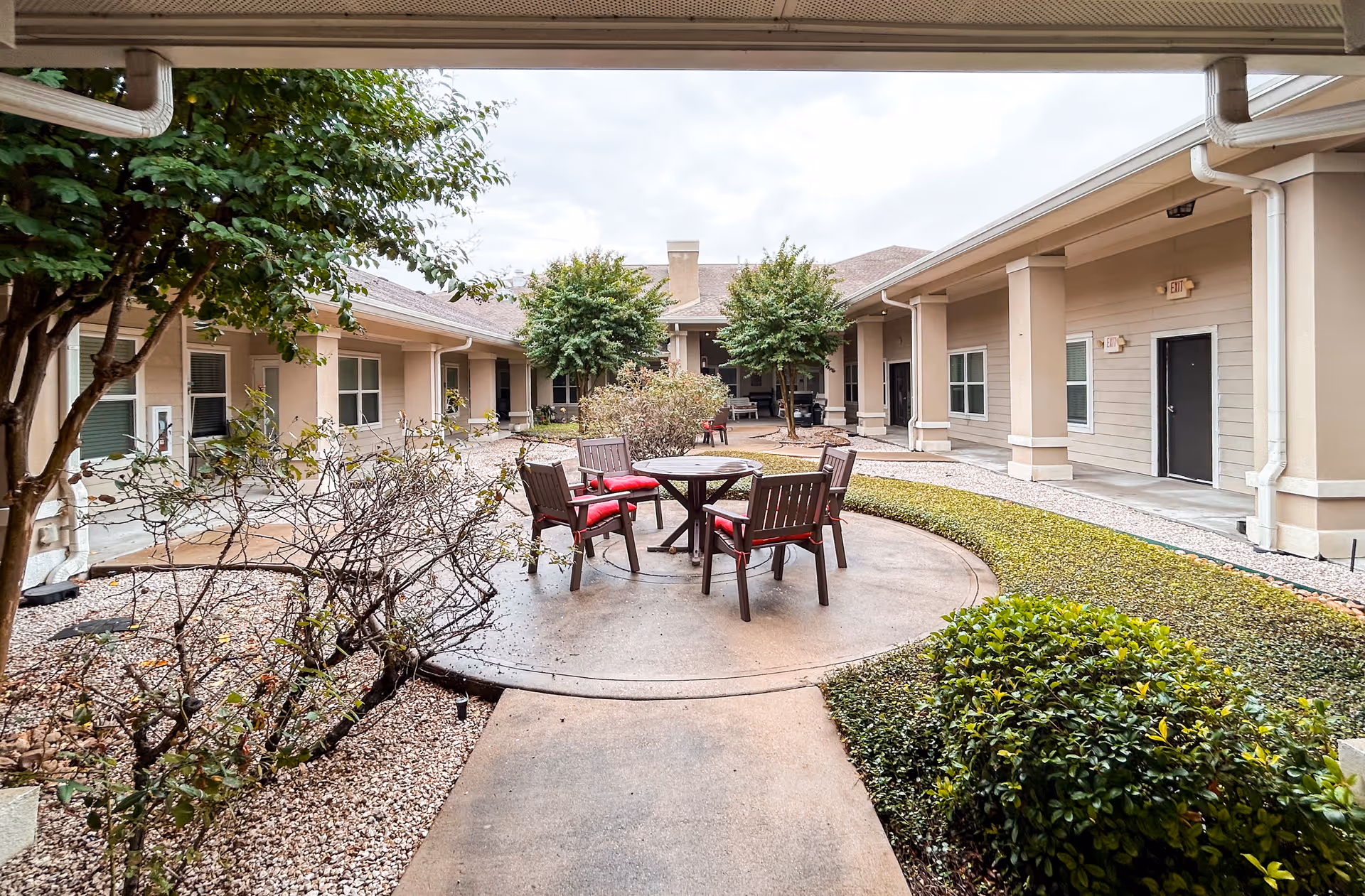 A central courtyard at a senior living facility with a round patio table and chairs, landscaping, and surrounding single-story building walkways.