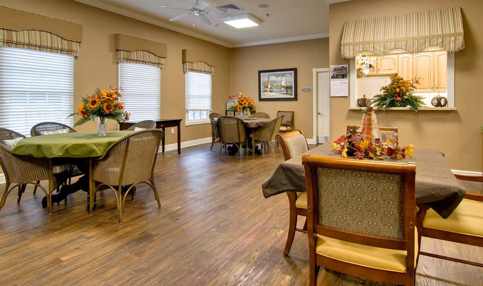 A cozy dining room with several tables covered with tablecloths and decorated with flower arrangements. The room has wooden flooring, beige walls, three large windows with blinds, and a ceiling fan. There is a pass-through window to a kitchen area with cabinets and more floral decorations.