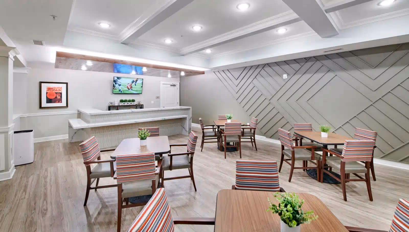 A bright and modern dining area with several wooden tables and chairs featuring striped upholstery. Each table has a small potted plant as a centerpiece. The room has light wood flooring, a decorative wall with geometric patterns, and a white ceiling with recessed lighting. A TV is mounted on the wall above a counter with a marble finish, and a framed picture is hung on the adjacent wall.