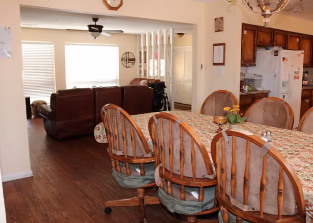 Open-plan dining area with a long table and wooden chairs, looking toward a living room with brown sofas and a kitchen in the background.