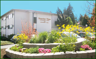 Front exterior of Sunrise View retirement facility with colorful curved flower beds and parked cars.