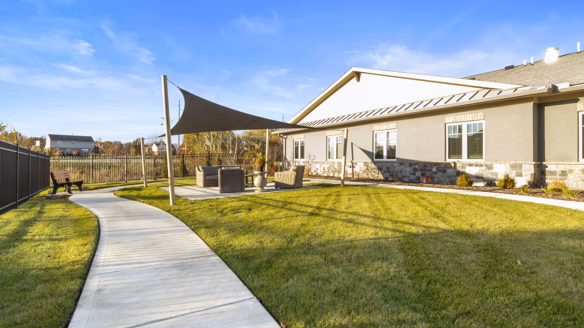 Sunlit outdoor courtyard with a curved concrete path, grassy lawn, shaded seating area and a low building in the background.