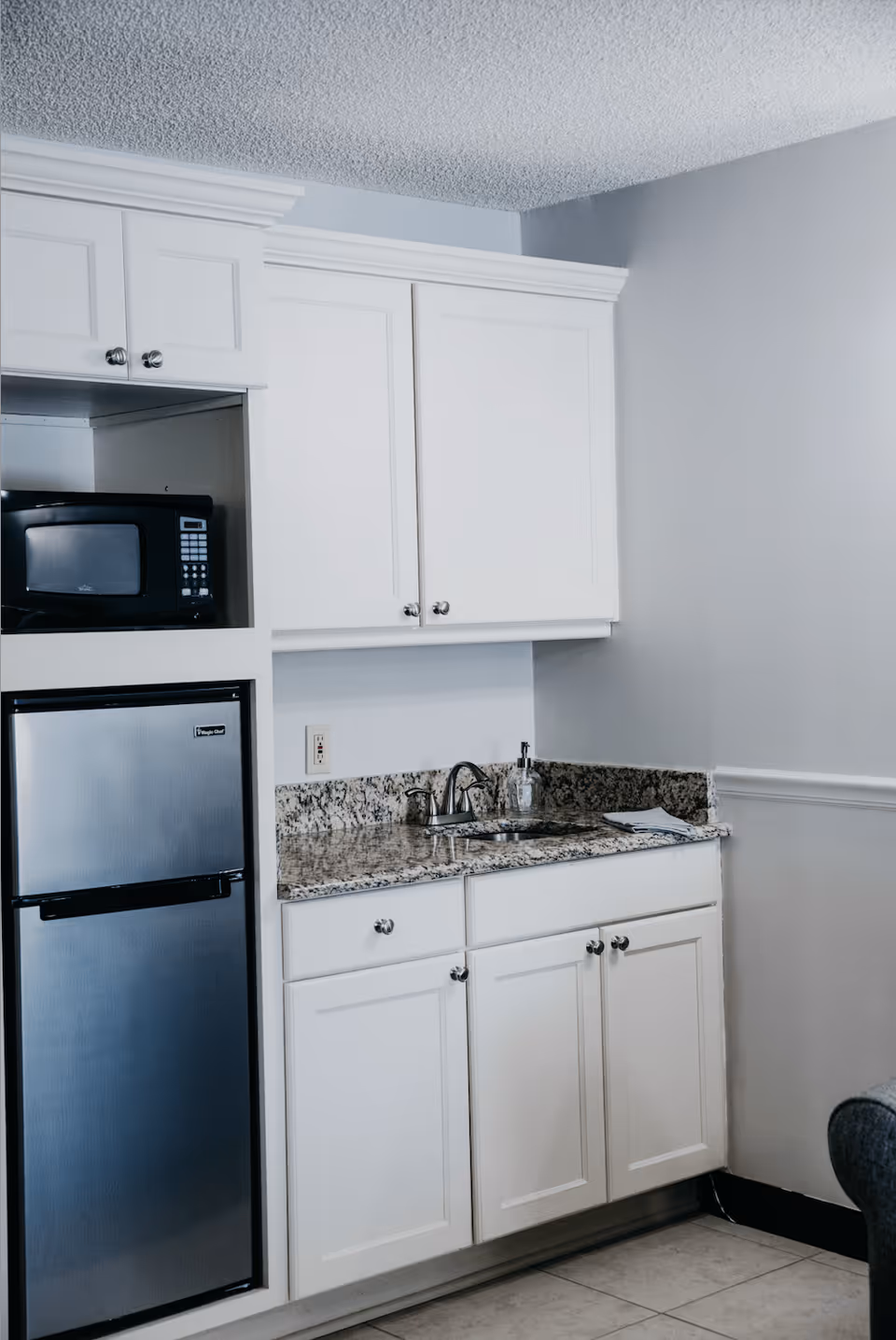 A small kitchen area with white cabinets, a granite countertop, a stainless steel mini refrigerator, a black microwave, a sink with a faucet, and a soap dispenser. The walls are painted light gray and the floor is tiled.